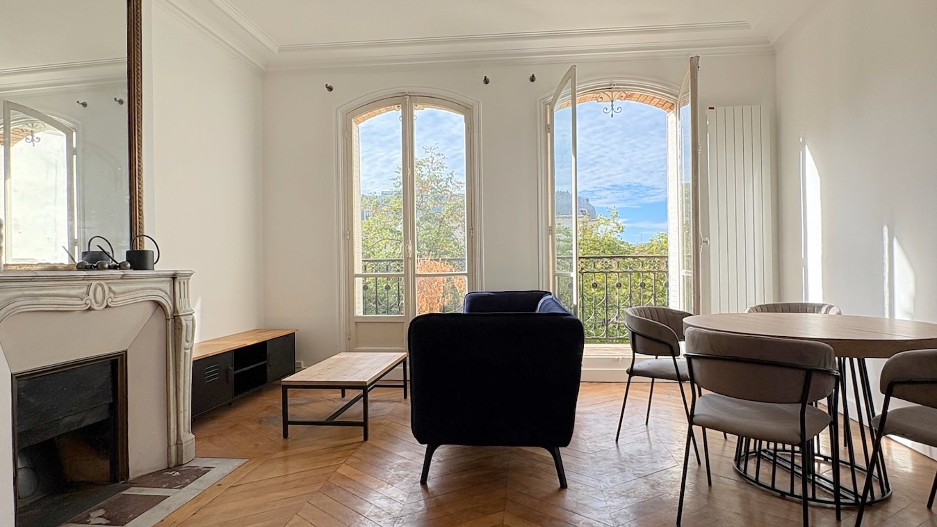 Séjour haussmannien à Paris : canapé en velours bleu nuit, table ronde en bois, cheminée en marbre et parquet en chevrons